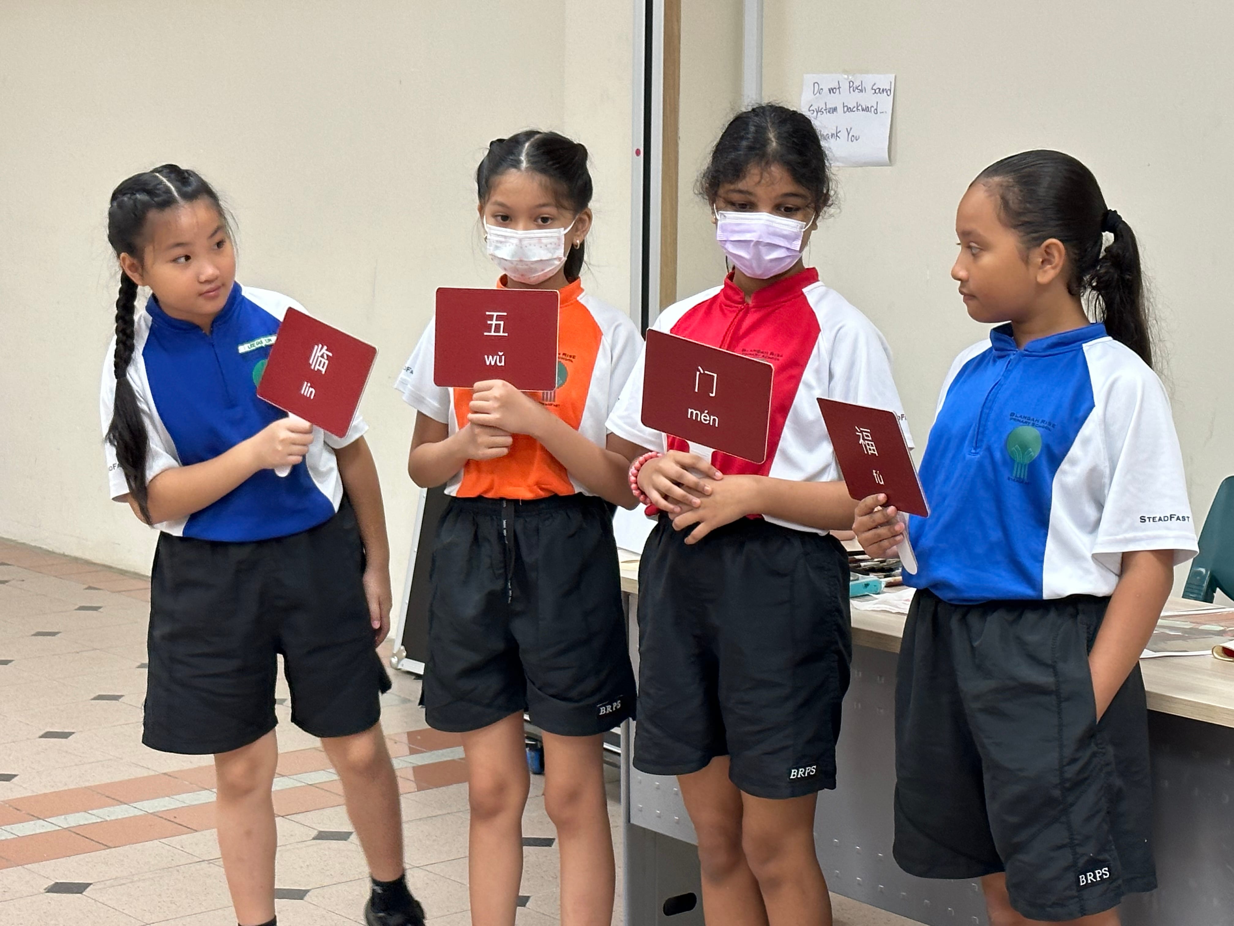Four school girls in uniforms hold red cards displaying Chinese characters with pinyin romanization.
